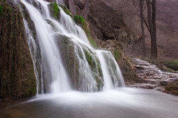 Fototapeta premium Long exposure view of a sunlit waterfall with soft streams of water falling down red rocks covered by green moss