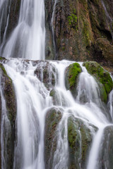 Fototapeta premium Close up, long exposure view of a sunlit waterfall with soft streams of water falling down red rocks covered by green moss