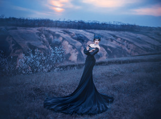 grim lady with pale white skin and dark hair beside flowering tree, witch turns with black crow with long luxurious dress and open shoulders, gothic image and makeup, cold metal crown and jewels