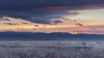 Distant Pirot city covered by mist and beautiful colors of sunset sky and silhouette horizon mountains panorama