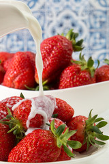 Close up. Someone pours milk (cream) into a plate of delicious juicy strawberries. Blue and white background.
