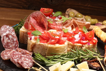 Traditional italian antipasto bruschetta appetizer with cherry tomatoes, cream cheese, basil leaves and balsamic vinegar on cutting board with prosciutto, salami, cheese