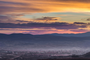 Dramatic, colorful sunset above distant city covered in mist and silhouette horizon mountains