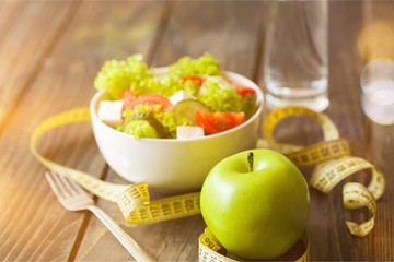 Salad in white bowl, fresh green apple and tape measure on dark wooden background. Diet and weight loss concept. - Powered by Adobe