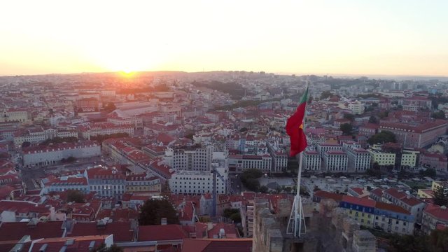 Aerial view medieval fortress on hill top in Lisbon. Drone flying above moorish Castelo de Sao Jorge near flapping poruguese flag on the wall and people enjoying picturesque cityscape at sunset