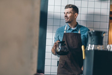 Smiling barista with coffee jug standing with his back to the wall