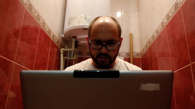 Bearded man typing on a laptop while sitting on the toilet.