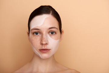 Beautiful young woman with white facial mask standing against beige background