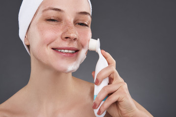 Obraz premium Smiling young woman with freckles using pore cleansing brush after bath