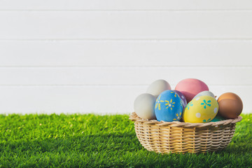 Easter eggs handmade in the basket on green grass with white wood wall background.