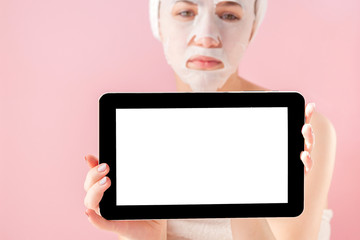 Beautiful girl with a tissue mask and a tablet in their hands with copy space on a pink background. Healthcare and beauty treatment and technology concept