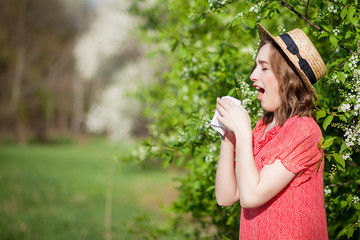 Young girl blowing nose and sneezing in tissue in front of blooming tree. Seasonal allergens affecting people. Beautiful lady has rhinitis.