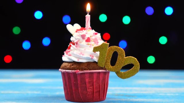 Delicious birthday cupcake with burning candle and number 100 on multicolored blurred lights background