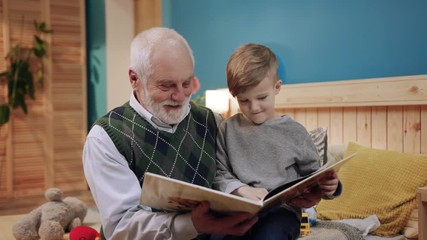 Happy grandfather and grandson while reading a funny book sitting at home generation book happiness child reading kid man caucasian family childhood grandchild slow motion - Powered by Adobe