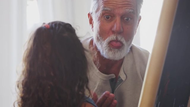 Senior man joking with his granddaughter while she draws on a blackboard, close up