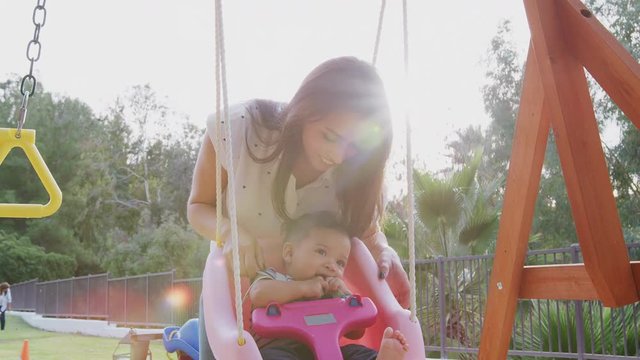 Young Hispanic mother pushing her baby on a swing at a playground in the park, close up