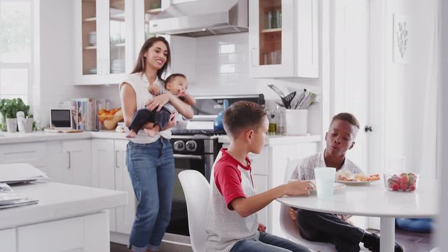 Mother holding baby stands in the kitchen talking with her son and his friend, over for a playdate