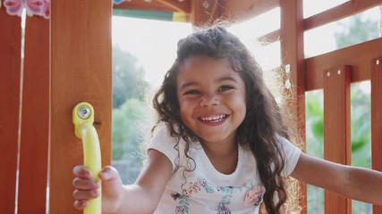 Young Hispanic girl playing on a climbing frame in a playground smiling to camera, backlit, close up