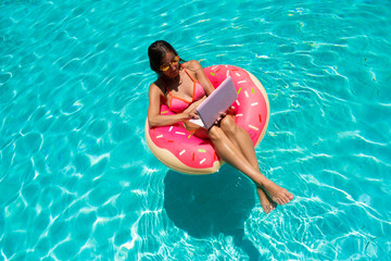 Young female freelancer sitting with a tablet in an inflatable circle in the pool. Aerial view