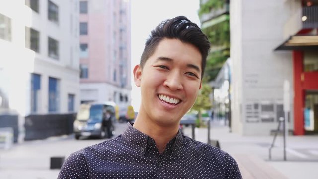 Millennial Asian man sitting in a city street laughing to camera, head and shoulders close up