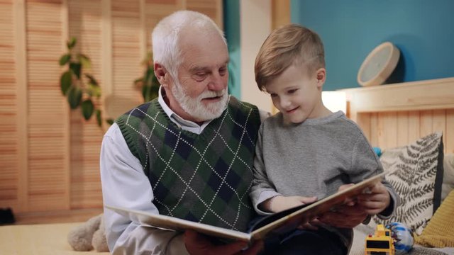 Old man with gray hair and cute grandson reading illustrated book with fairytales.  Family members spending unforgettable time together. Indoors. Happy childhood.