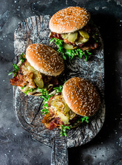 Classic homemade burger with pork cutlet, bacon, pickles, fried onions and mayonnaise mustard sauce on wooden chopping board, on dark background