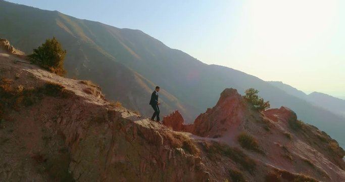 Young confident businessman wearing suit and tie going up the rocky mountain, representing way to success - 4k aerial shot