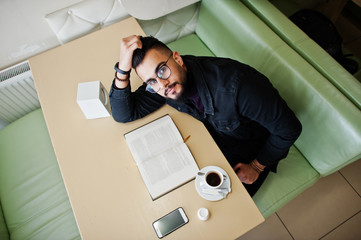 Arab man wear on black jeans jacket and eyeglasses sitting in cafe, read book and drink coffee....