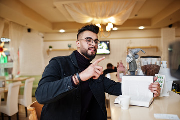 Fototapeta premium Arab man wear on black jeans jacket and eyeglasses in cafe drink coffee at bar with book. Stylish and fashionable arabian model guy.