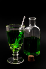 Fototapeta na wymiar Alcoholic drink, creative stimulant and bohemian lifestyle concept theme with glass of green absinthe and stainless steel spoon next to a vintage bottle and a corck isolated on black background