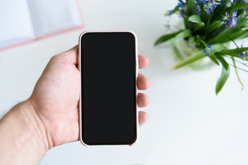 Male hand with a smartphone. Black blank screen. Table with notebook and flowers on background