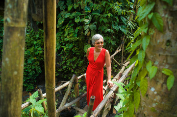 Naklejka na ściany i meble natural lifestyle portrait of beautiful and happy middle aged 40s or 50s Asian woman with grey hair in stylish red dress walking on green tropical jungle landscape