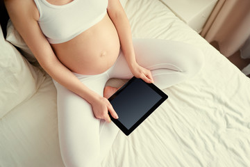 App for women. Copy space on screen. Top view of young pregnant woman holding tablet computer sitting on a bed at home.