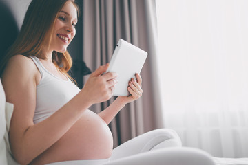 Technology and pregnancy. Young pregnant  woman using tablet computer sitting on a bed at home.