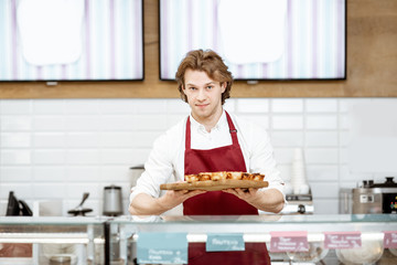 Portrait of a handsome salesman or confectioner in red apron standing with fresh baked pastel de nata in the pastry shop