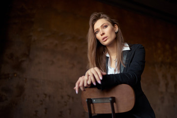 Fototapeta na wymiar Studio portrait of young beautiful sensual woman in blak suit sitting on wooden chair against dark background.