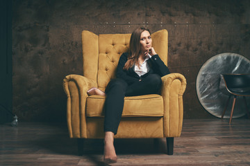 Beautiful young stylish woman in black suit sitting in yellow armchair.