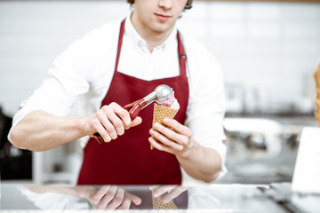 Handsome salesman in red apron making ice cream in waffle cone for selling in the modern pastry shop