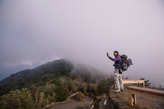 Hiker with backpack staying on mountain top above clouds and jungle valley, doing pictures or try to catch 3g internet on mobile phone