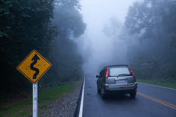 SUV car parking near the bullet riddled on the curvy road sign on the misty road.