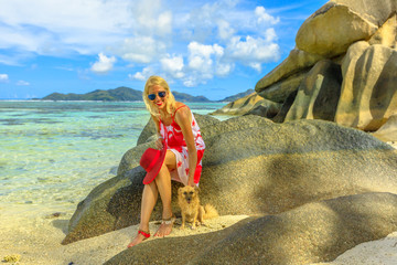 Smiling lifestyle woman sitting on a boulders, touches a cute dog at Anse Source d'Argent one of the most beautiful beaches in La Digue, Seychelles famous for crystal sea and large granite rocks.