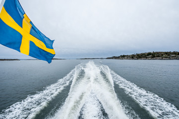 Aft view from a fast ferry cruising through calm waters on a grey day. The swedish flag waving from the ship.