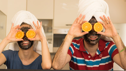 Sweet Orange Glasses of Happiness. Happy Smiling Couple - Man and Woman - Have Fun with Cosmetic Black Mud Mask on Their Faces and Orange Slices on Eyes. Skincare and Kitchen Humor Story, Concept.