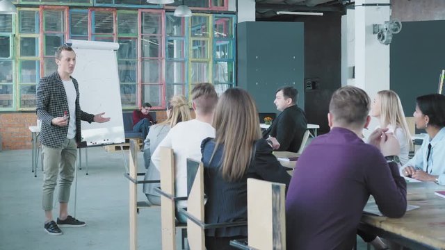 Start up business team holds a meeting at a large table. The man manager emotionally conducts a presentation near the flipchart and communicates with colleagues. Coworking. Creative office interior