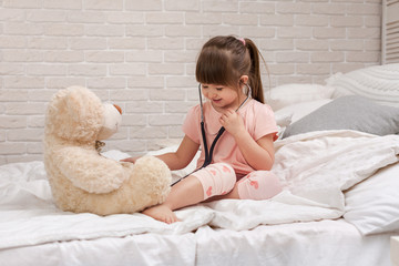 cute child girl playing doctor with teddy bear at home. girl listens a stethoscope to toy.