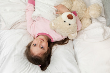cute little child girl in pyjamas with teddy bear in bed. good morning. top view. girl looking at camera