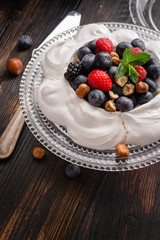Pavlova cake with fresh summer berries and hazelnuts on glass standcake  on black wooden table.