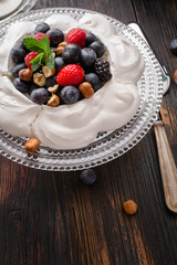 Pavlova cake with fresh summer berries and hazelnuts on glass standcake  on black wooden table.