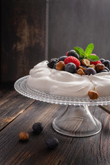 Pavlova cake with fresh summer berries and hazelnuts on glass standcake  on black wooden table.
