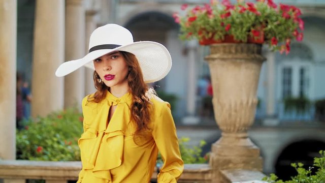 Young beautiful fashionable lady wearing vintage stylish yellow blouse, white wide brim hat, earrings, posing in old european city
